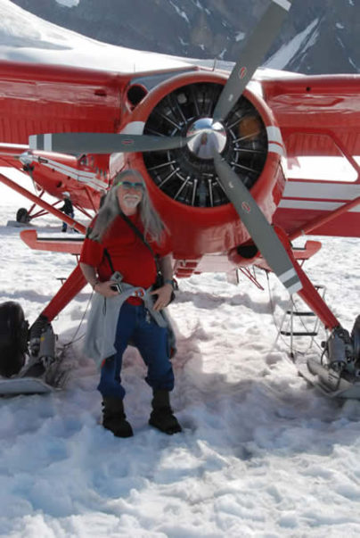Silver haired Norman Greenbaum stands on snow in front of propeller of red airplane.