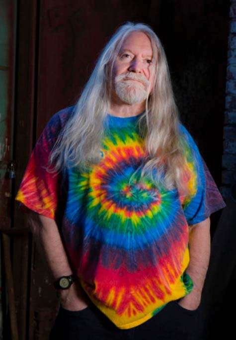 Silver-haired Norman Greenbaum wearing a spiral tie dye shirt. His hands are in his pockets.